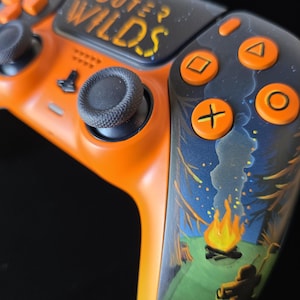May include: Custom orange and black video game controller with the text "OUTER WILDS". The controller features a forest scene with a campfire. The buttons are orange, and the joysticks are black.