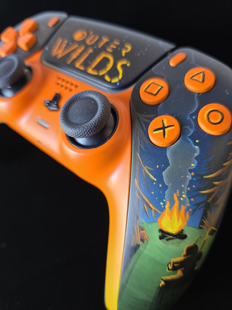 May include: Custom orange and black video game controller with the text "OUTER WILDS". The controller features a forest scene with a campfire. The buttons are orange, and the joysticks are black.