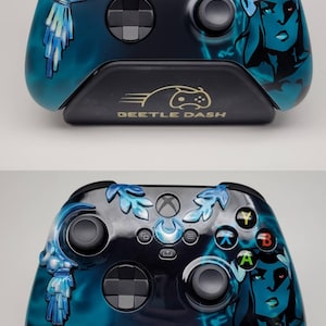 May include: Custom Xbox controller with a blue and black design. The controller features a matte finish and a stylized illustration of a woman's face on the right side. The controller rests on a black stand with the text "BEETLE DASH".