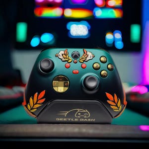 Könnte beinhalten: Benutzerdefinierter Xbox-Controller in Teal und Schwarz mit goldenen Akzenten. Der Controller hat ein dekoratives Design mit Lorbeerblättern und dem Text "BEETLE DASH" auf der Unterseite. Die Knöpfe sind gold und rot.