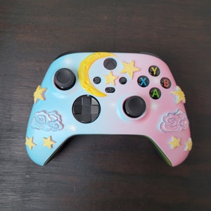 Könnte beinhalten: Ein individuell lackierter Videospiel-Controller mit einem pastellblauen und rosa Farbverlauf. Der Controller hat einen gelben Mond, Sterne und Wolken. Die Knöpfe sind schwarz und die Funktionstasten sind farbig.