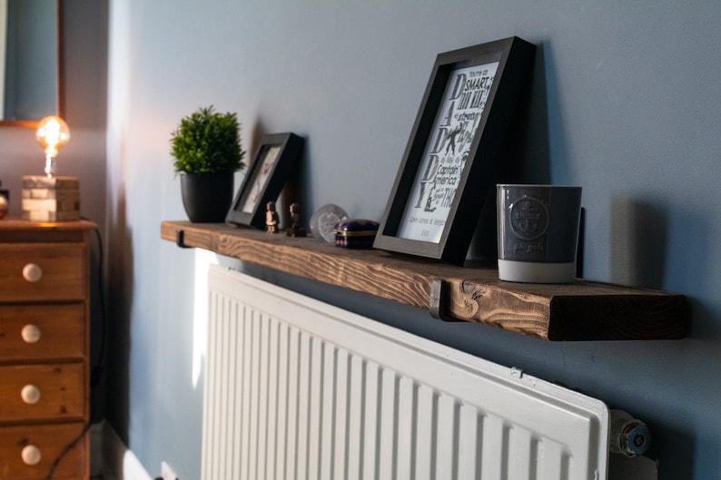 May include: A rustic wooden shelf with black metal brackets is mounted above a white radiator. The shelf is decorated with a potted plant, two framed pictures, and a candle. The picture on the right has the text "Daddy" in large letters.