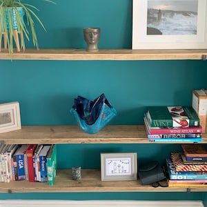 May include: Three wooden shelves with metal brackets against a teal wall. The top shelf has a small ceramic sculpture of a face, a potted plant, and a framed photo. The middle shelf has a blue glass sculpture and a stack of books. The bottom shelf has a row of books, a small framed photo, a black purse, and a stack of books.