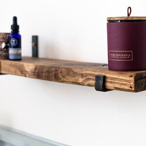 May include: A rustic wooden shelf with metal brackets, holding a burgundy candle with a gold label that reads "Home Fragrance Scented Candle" and a small blue bottle with a dropper.