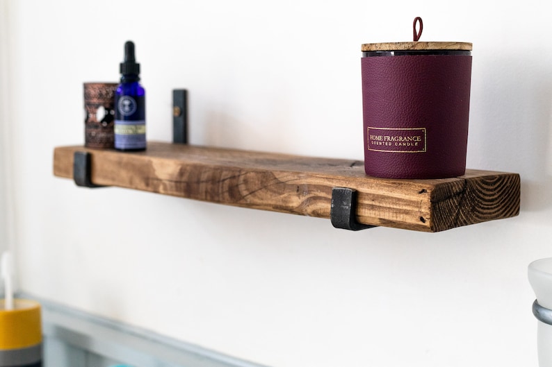 May include: A rustic wooden shelf with metal brackets, holding a burgundy candle with a gold label that reads "Home Fragrance Scented Candle" and a small blue bottle with a dropper.