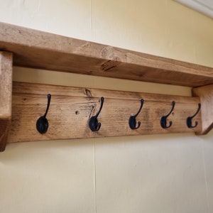 May include: A wall-mounted coat rack crafted from light brown wood with a natural grain finish. The unit features a flat top shelf supported by two side brackets and a horizontal back panel equipped with five evenly spaced black metal hooks.