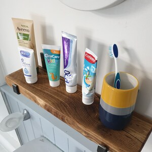 May include: A wooden shelf with four tubes of toothpaste and a toothbrush in a yellow, gray, and blue ceramic holder. The toothpaste brands are Aveeno, OraCare Kids, Pro, and Colgate. The shelf is mounted on a white wall.