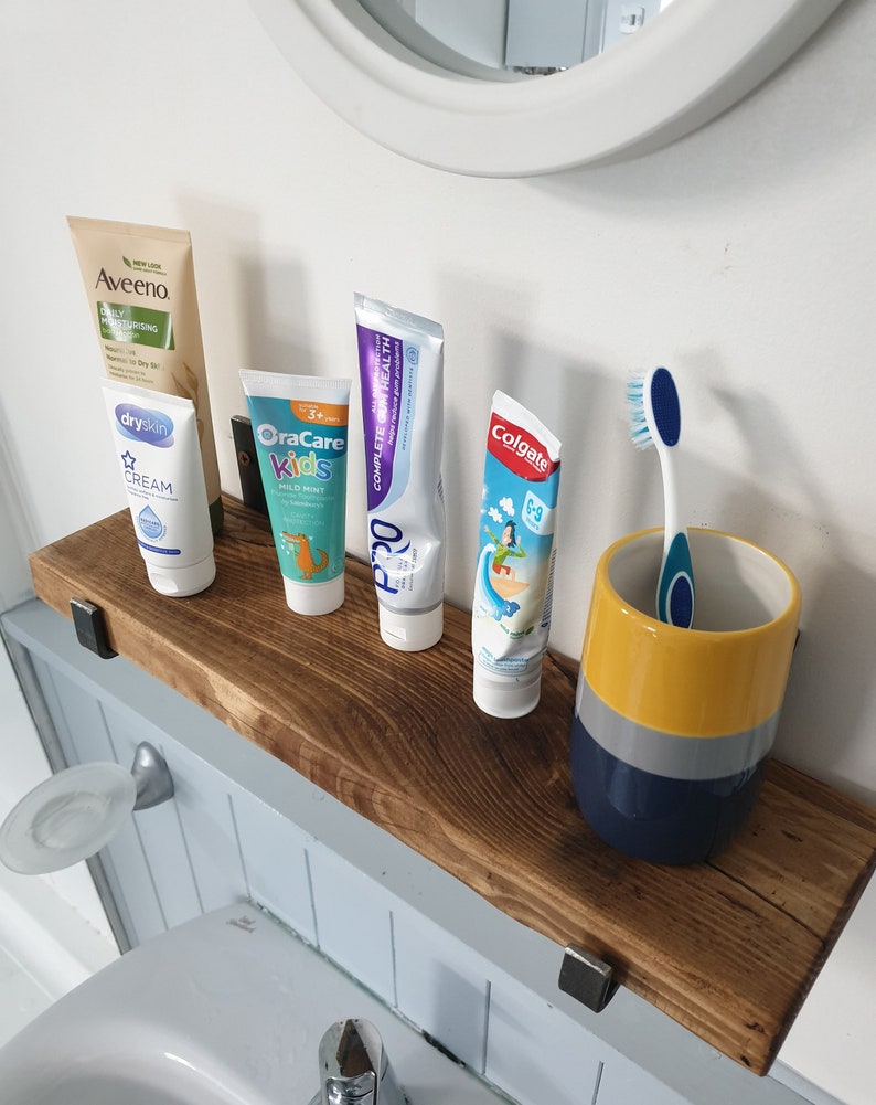 May include: A wooden shelf with four tubes of toothpaste and a toothbrush in a yellow, gray, and blue ceramic holder. The toothpaste brands are Aveeno, OraCare Kids, Pro, and Colgate. The shelf is mounted on a white wall.