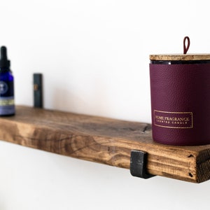 May include: A wooden shelf with metal brackets, holding a small bottle of essential oil, a decorative candle with a burgundy leather-like exterior and a wooden lid, and a small decorative object. The candle has a gold label that reads "Home Fragrance Scented Candle".