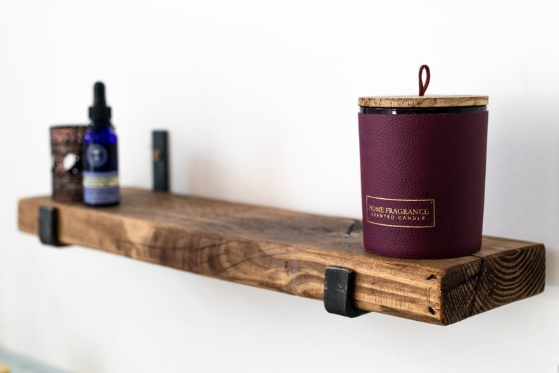 May include: A wooden shelf with metal brackets, holding a small bottle of essential oil, a decorative candle with a burgundy leather-like exterior and a wooden lid, and a small decorative object. The candle has a gold label that reads "Home Fragrance Scented Candle".