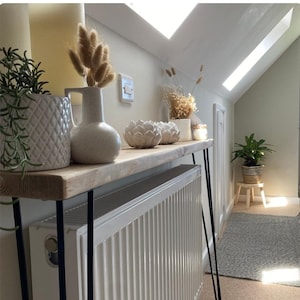 May include: A white radiator with a wooden shelf above it. The shelf is decorated with dried flowers, a white ceramic vase, and white ceramic decorative items. The shelf is supported by two black metal legs.