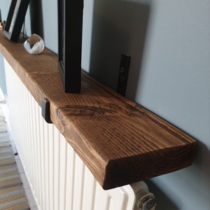 May include: A dark brown wooden shelf mounted above a white radiator. The shelf is supported by two black metal brackets. The shelf has a natural wood grain and is stained with a dark brown finish.