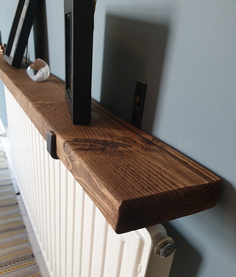 May include: A dark brown wooden shelf mounted above a white radiator. The shelf is supported by two black metal brackets. The shelf has a natural wood grain and is stained with a dark brown finish.