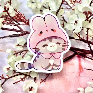 May include: A sticker of a cartoon cat wearing a pink bunny hat. The cat has a striped tail and is sitting with its paws tucked under its body. The sticker has a white outline.