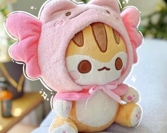Puede incluir: Un juguete de peluche de gato con un disfraz de rana con capucha rosa. El gato tiene ojos marrones, una nariz marrón y una corbata de lazo rosa. La capucha de rana tiene un lazo rosa en la parte posterior.