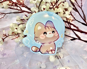 May include: A sticker featuring a cartoon cat with brown and white fur, enclosed within a blue bubble. The cat has large eyes and a pink nose. The sticker is positioned on a white background adorned with a white, sparkly, star-shaped fabric.
