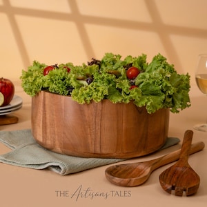 May include: A large wooden salad bowl filled with fresh lettuce, tomatoes, and other salad ingredients. Two wooden serving spoons and a fork rest beside the bowl on a light green napkin. The Artisan's Tales logo is at the bottom.