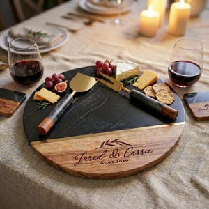a round wooden platter with a black marble surface, displaying a selection of cheese, crackers, and grapes. The platter is accompanied by two wine glasses filled with red wine, and a set of serving utensils, including a spatula and tongs, arranged on the platter. The image also includes a personalized inscription that reads "Jared & Cassie" and "01.06.2020".