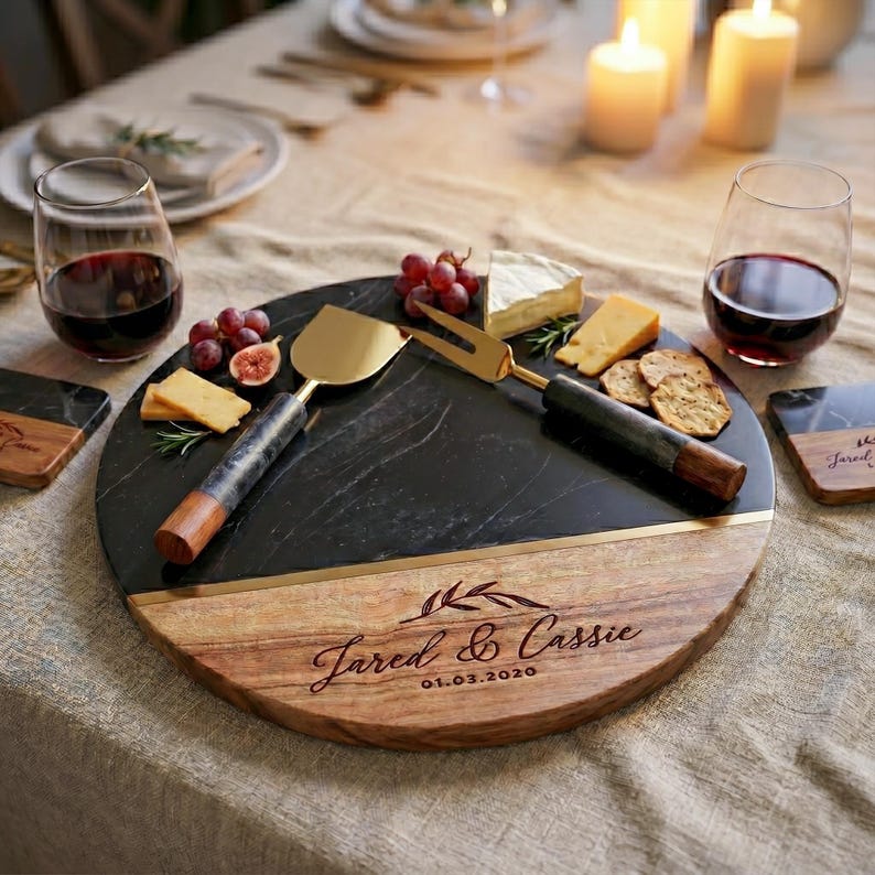 a round wooden platter with a black marble surface, displaying a selection of cheese, crackers, and grapes. The platter is accompanied by two wine glasses filled with red wine, and a set of serving utensils, including a spatula and tongs, arranged on the platter. The image also includes a personalized inscription that reads "Jared & Cassie" and "01.06.2020".