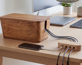 May include: A rectangular wooden cable management box with a removable lid, featuring a side slot for cords. A matching wooden multi-port charging hub is connected to the box, holding several charging cables and a smartphone on the desk surface.