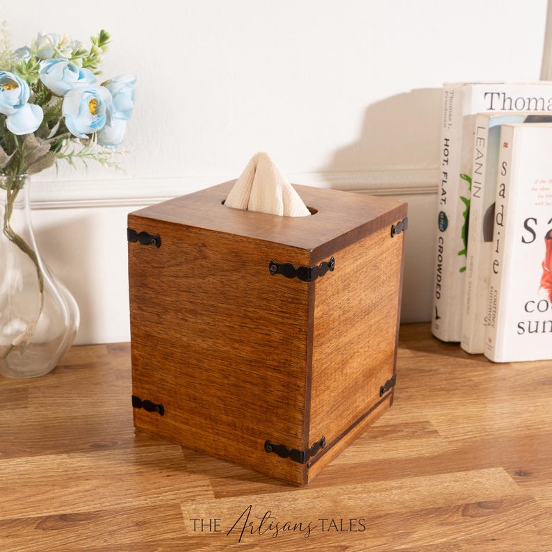 A wooden tissue box with a white napkin inside sits on a wooden surface, accompanied by a vase of blue flowers and a stack of books.