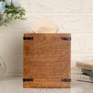 A wooden tissue box with a white cloth napkin inside, placed on a white countertop next to a stack of books.