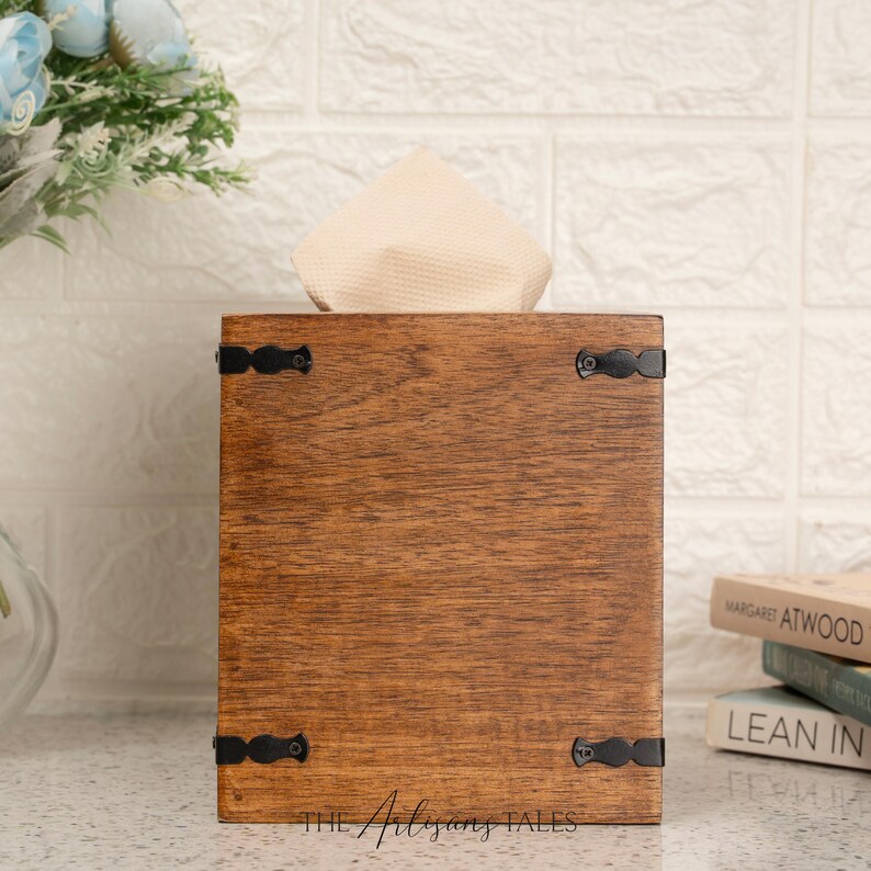 A wooden tissue box with a white cloth napkin inside, placed on a white countertop next to a stack of books.