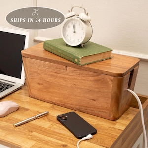 May include: A rectangular wooden cable management box with a lid, featuring a side cutout for cords. A green hardcover book and a white analog alarm clock sit on top of the box. A black smartphone with a white charging cable is placed in front.