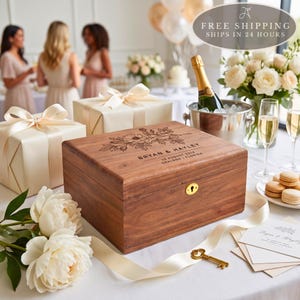 May include: A rectangular wooden keepsake box with a hinged lid featuring a laser-engraved floral design and the text BRYAN & HAYLEY 25 AUGUST 2026 ORLANDO FLORIDA. The box has a gold-colored keyhole on the front and sits on a white surface.
