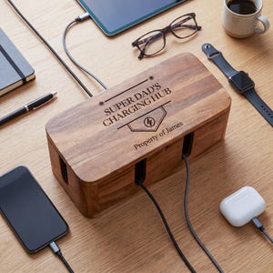 May include: A wooden charging hub with the text "SUPER DAD'S CHARGING HUB" and "Property of James". The hub has slots for charging devices, with cables connected to a phone, tablet, and earbuds. Other items include glasses, a watch, and a coffee cup.