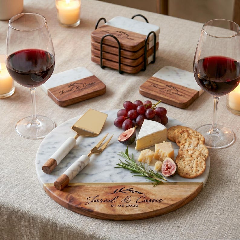 a table setting with a marble platter containing cheese, crackers, grapes, and a knife, accompanied by two glasses of red wine. The table also has a set of coasters and a small tray with additional cheese and crackers.