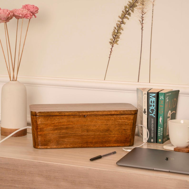 a wooden box, a laptop, a vase with pink flowers, and a stack of books on a desk.