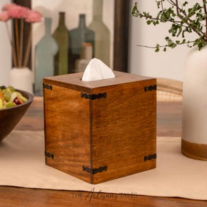 A wooden tissue box with a white napkin in it, placed on a wooden table. In the background, there are several bottles and a plant.