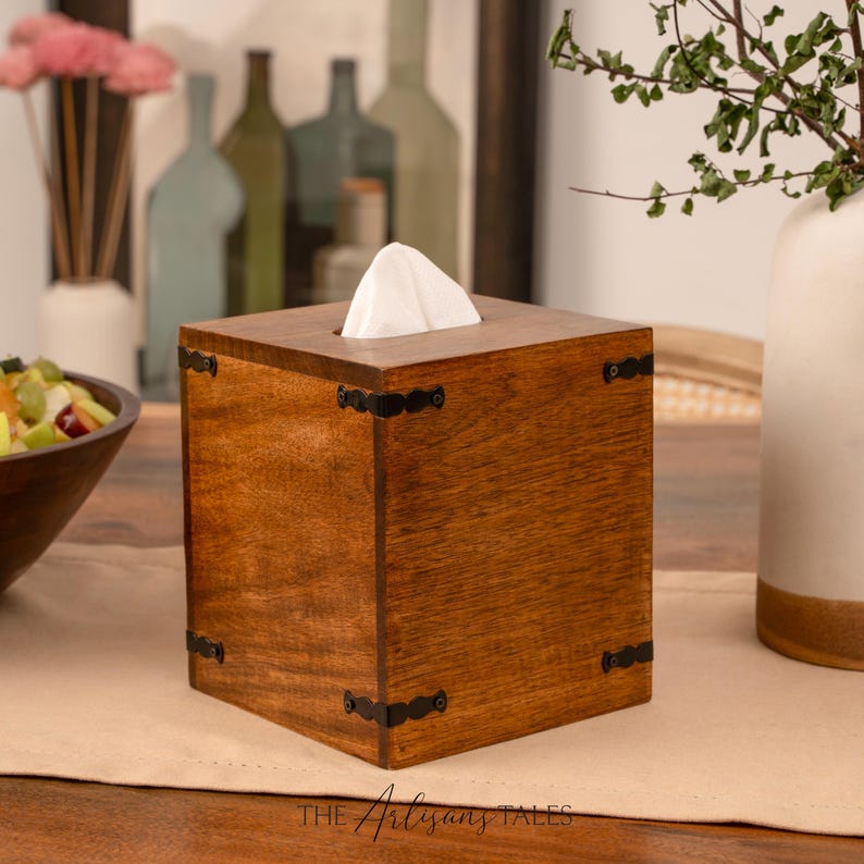 A wooden tissue box with a white napkin in it, placed on a wooden table. In the background, there are several bottles and a plant.