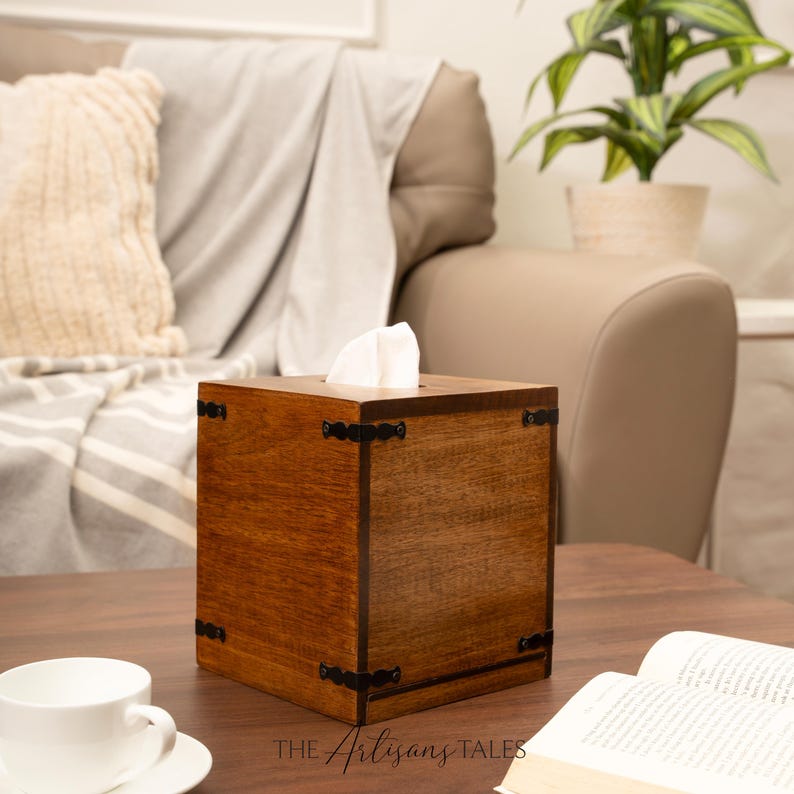 A wooden tissue box with a white tissue on top, placed on a wooden coffee table in a cozy living room setting with a beige couch and a potted plant in the background.