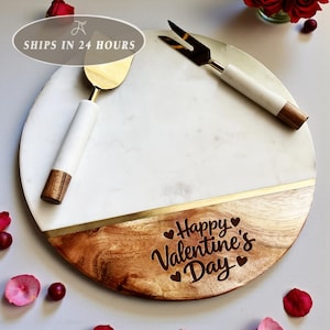 Puede incluir: Una tabla redonda para servir de mármol y madera con detalles dorados. Incluye un cuchillo y un tenedor para queso con mangos blancos y hojas doradas. La tabla está grabada con "Happy Valentine's Day."