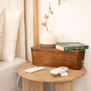 a wooden nightstand with a vase of flowers, a book, a remote control, and a smartphone charging on it.