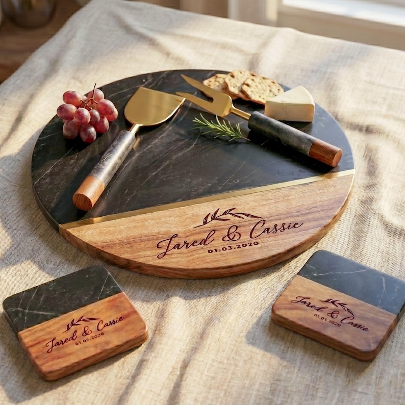 a round wooden platter with a black marble surface, containing a variety of food items such as grapes, cheese, crackers, and a knife. The platter is accompanied by two coasters, each with the name "Jared & Cassie" printed on them.