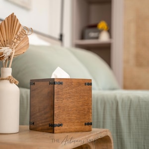 A wooden tissue box with a white tissue paper inside sits on a wooden table, accompanied by a white vase with dried flowers and a green pillow in the background.