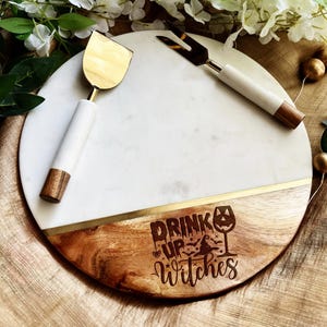 May include: A round serving board made of white marble and wood with a gold metal accent strip. The wood section features the engraved text "DRINK UP Witches" with a wine glass, witch hat, and bat icons. Includes a gold cheese knife and fork.