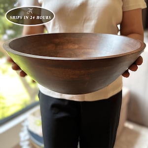 May include: A large, round, dark brown wooden bowl with a smooth, matte finish and a wide, open top. The bowl features a deep, curved basin and a thick, solid rim, showcasing the natural grain patterns of the wood throughout its exterior surface.