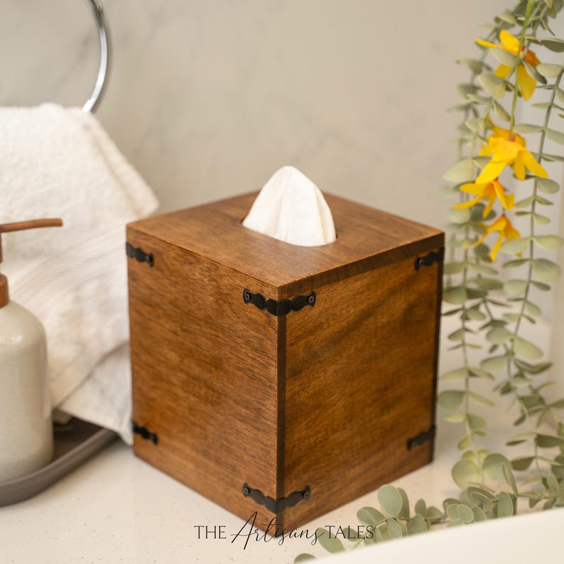 A wooden tissue box with a white tissue paper, placed on a white countertop next to a soap dispenser and a plant.