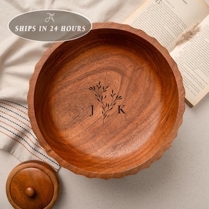 May include: A round wooden bowl with a scalloped edge and a natural brown wood grain finish. The center of the bowl features a dark engraved design of a leafy branch positioned between the capital letters J and K in a serif font.