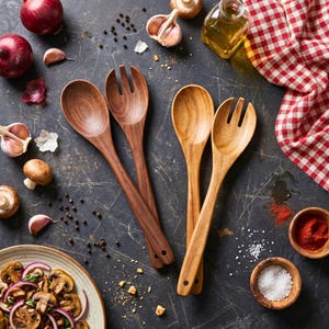 May include: Overhead shot of wooden salad spoons and forks, with a dark brown finish, alongside ingredients like red onions, garlic, mushrooms, and spices. A red and white checkered cloth is in the background.