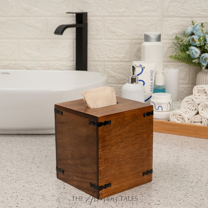 A wooden tissue box with a black metal frame sits on a bathroom counter, next to a white sink and various toiletries.