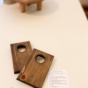 May include: Two wooden coin toss game boards with a hole in the center. The boards are brown and have a natural wood grain. The boards are on a white surface with two silver quarters. The text on the white card reads: "Coinhole Rules: Quarters must bounce off the surface in front of the board. 3 points for a quarter in the hole, 1 point for a quarter on the board. Alternate turns bouncing two quarters each. The scores cancel one another. Play to 21."