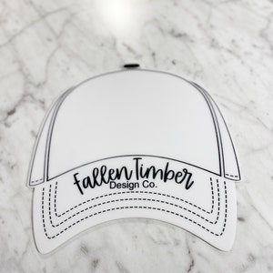 May include: White blank baseball cap with a black outline and the text "Fallen Timber Design Co." in black cursive lettering.