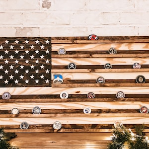 May include: A wooden American flag display with a dark brown stain. The flag is made of wood and has a distressed look. There are slots for displaying challenge coins or other small items. The flag is mounted on a white brick wall.