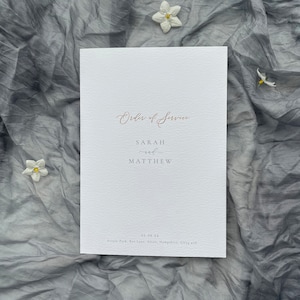 May include: White wedding ceremony order of service with the names Sarah and Matthew printed in a light grey font. The date 06.08.24 and the location Feople Park, Rye Lane, Alion, Hampshire, GU4 all are printed at the bottom of the page.