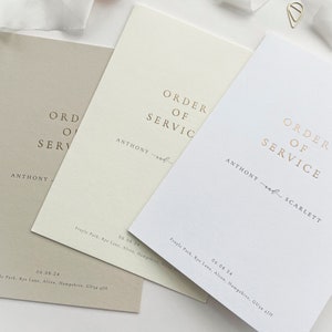 May include: Three white cards with gold lettering that reads "Order of Service" with the names Anthony and Scarlett. The cards are on a white surface with pink flower petals.